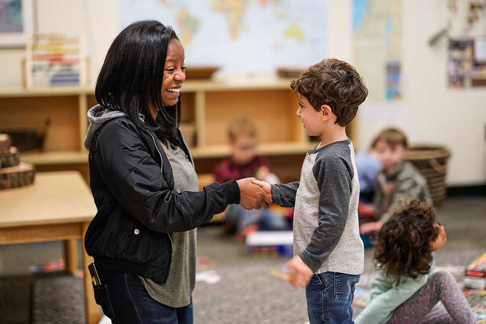 An ECP Preschool teacher smiles and shakes a boy's hand in a classroom.