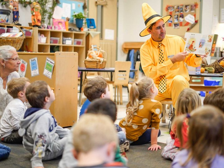 A man with a yellow hat reads a book to a group of preschool kids.