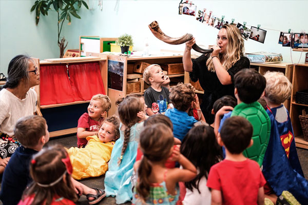 Preschool teacher blowing a shofar in front of kids in classroom