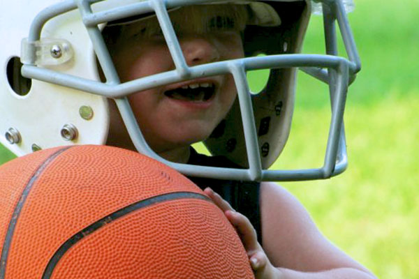 A toddler child wears a football helmet and holds a basketball outside on a grass field.