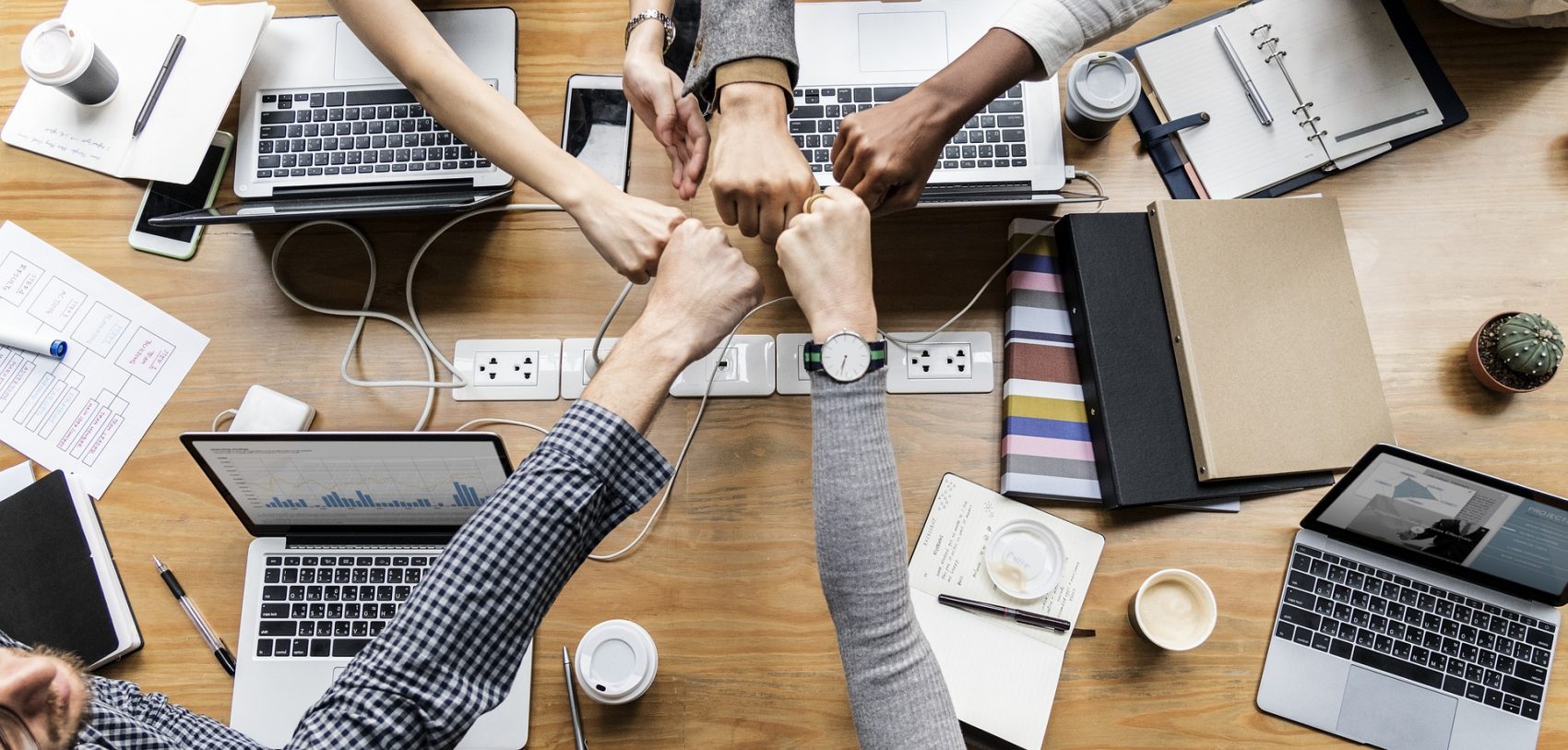 Group of people at a professional meeting put fists together over a table with laptops.