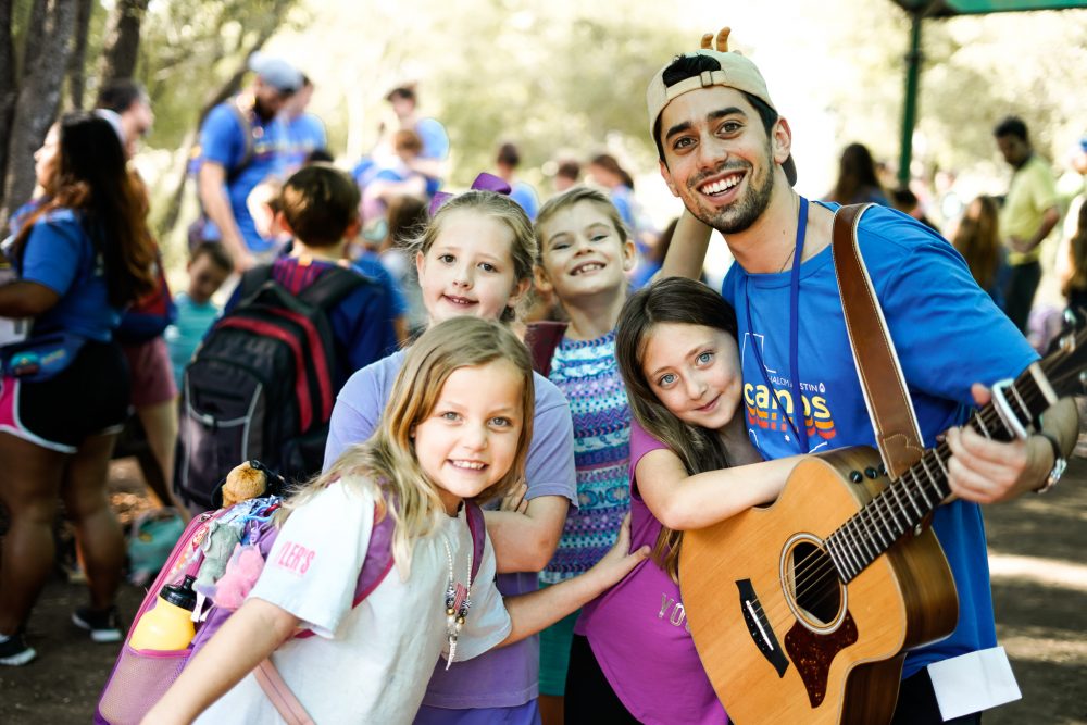 Four school-age girls smile with Mr. Max as he holds his guitar outside at JCamps