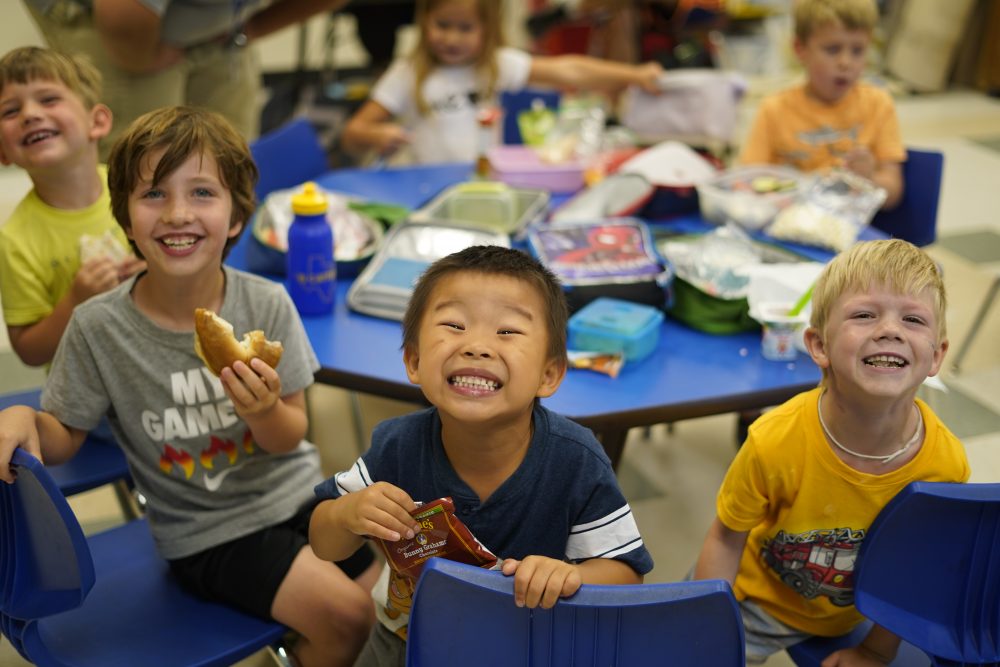 A group of kids sitting at a blue table smile during lunch or snack at JCamps.