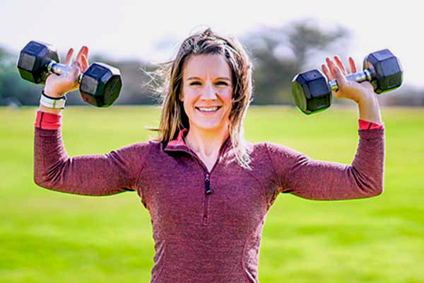 Personal trainer Chelsea Hanson lifts weights with her arms outside in a grass field.